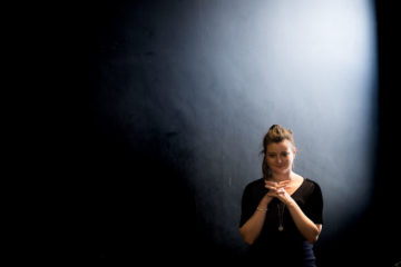 Sinead Costelloe in rehearsal. A young woman stands against a black wall, her fingers interlaced