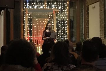 A photo of a young woman at an event reading in front of a microphone.